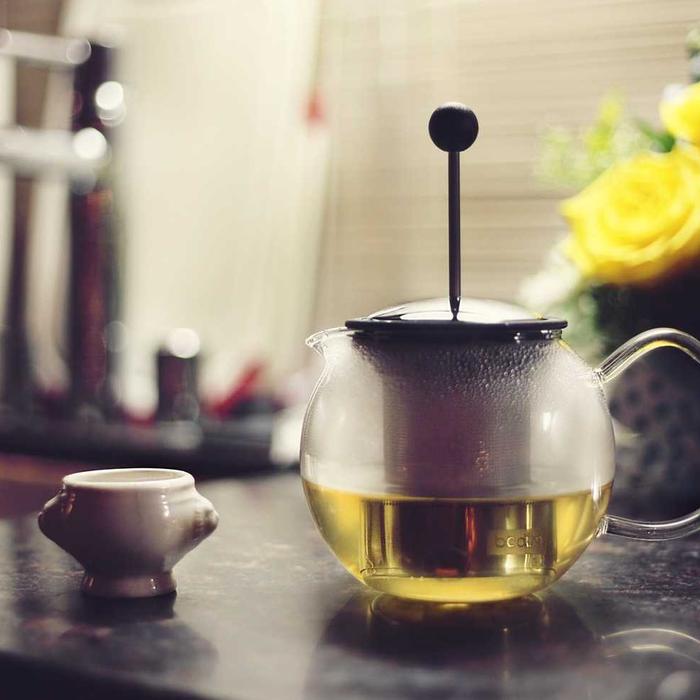 Glass teapot of golden tea beside a cup and yellow flowers
