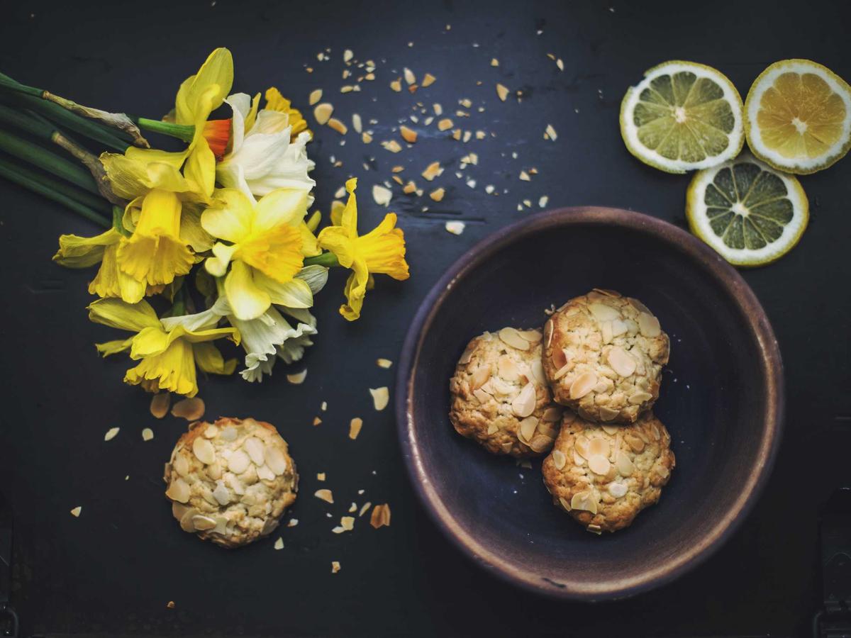 Golden almond biscuits in a dark bowl with lemon slices and daffodils on a dark table