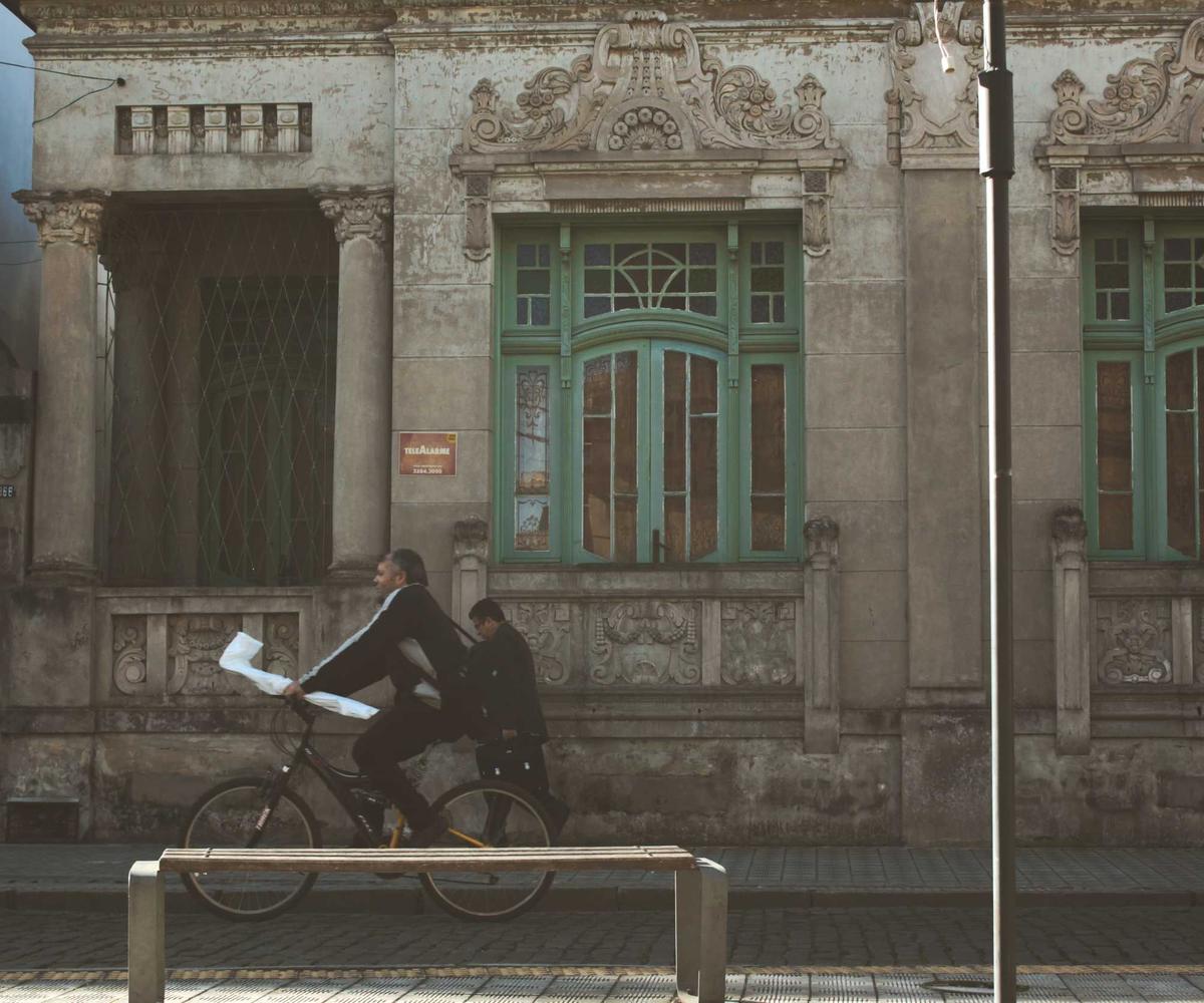 An old building with ornate plasterwork and tall teal-green window frames, a cyclist passing on the street below