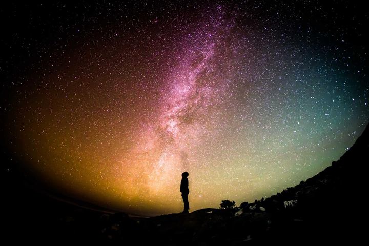 A person silhouetted on a ridge beneath a vivid pink and gold Milky Way